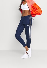 Kvinna iförd marinblå Adidas träningsoverallbyxor med vita ränder, vit sport-bh, orange jacka och vita sneakers, stående mot en neutral bakgrund.