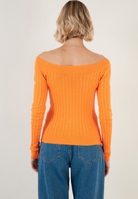 Pulover portocaliu cu umerii la vedere, cu maneci lungi și design ajustat. Textură tricotată ușoară. Asortat cu blugi cu talie înaltă, de culoare albastră.