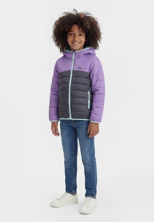 Lokkis juustega laps, kes kannab lilla ja halli kapuutsiga pufferjopet, siniseid teksaseid ja valgeid tossusid, naeratades ja seistes.