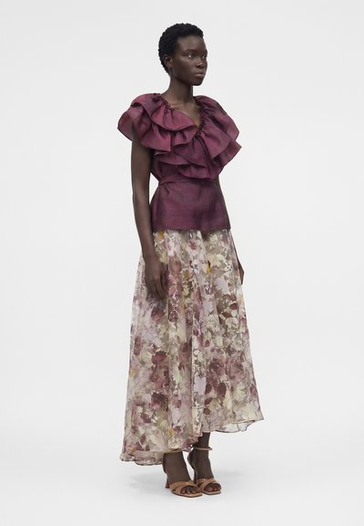 Γυναίκα που φοράει μωβ αμάνικο τοπ με βολάν και ζώνη και μακριά διάφανη φλοράλ φούστα, στέκεται με ουδέτερη έκφραση και μαυρισμένα σανδάλια.