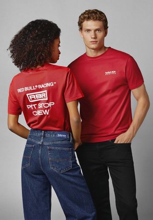 Rode T-shirts met korte mouwen met de tekst "RED BULL RACING" en "PIT STOP CREW". Blauwe denim jeans met achterzakken. Gladde texturen; eenvoudig ontwerp.