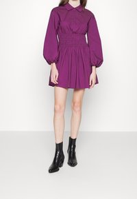 Robe en coton violet avec un col chemise, des manches bouffantes et une taille froncée. Portée avec des bottines noires et une jupe évasée.