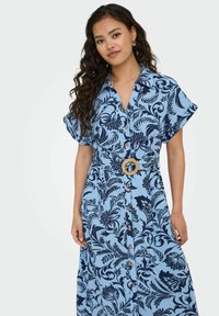 Hele sinine nööbidress, millel on tumesinine lillemuster, lühikesed rullitud varrukad, kokku tõmmatud vöökoht, millel on kudumivöö ja puidust nööpide aktsendid.
