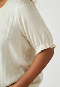 Hellbeige Bluse aus weichem, strukturiertem Stoff. Verfügt über gepuffte Ärmel mit gerafften elastischen Rüschen. Dezente vertikale Streifen setzen Akzente.