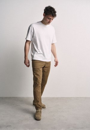 Ung mand iført en almindelig hvid T-shirt, brune bukser og beige sko, kigger ned, mens han står op ad en almindelig lys væg.