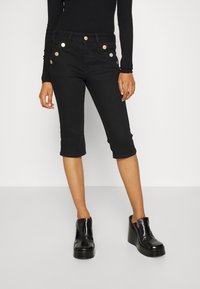 Pantalons capri en denim noir avec une coupe ajustée, dotés de boutons dorés et argentés le long des poches avant et d'un ourlet juste en dessous des genoux.