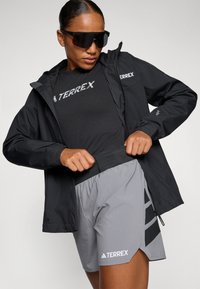 Svart lett jakke med hette, grå treningsshorts med svarte detaljer, og en svart t-skjorte med "TERREX" logo; mørke solbriller.