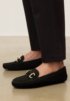 Czarne zamszowe mokasyny z złotym metalowym ozdobnikiem, noszone z czarnymi zwężanymi spodniami na beżowej podłodze.