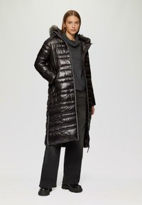 Langer schwarzer Puffercoat mit glänzender Oberfläche, gestepptem Design, abnehmbarem Kapuzenrand aus Fell und mit Reißverschlüssen versehenen Seitentaschen. Trägt man über einem grauen Pullover.