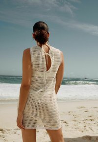 Vestido de ganchillo en color blanco roto con espalda descubierta, patrón de tejido abierto, borde de encaje en el dobladillo y escote atado. Fondo de playa visible.