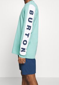 Měkké mintově zelené tričko s dlouhým rukávem a bílými rukávy, na kterém je navy modrý nápis "BURTON". V kombinaci s navy modrými šortkami. Materiál: bavlna.