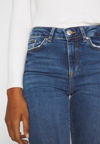 Calças de ganga azul de cintura alta com uma textura suave, costuras douradas e dois bolsos frontais com pequenos detalhes de desgaste.