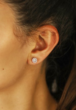 Runde Steckerohrringe mit einem zentralen weißen Stein, umgeben von einem Halo aus kleinen, klaren Schmucksteinen. Die Metallfassung ist in Silberoptik.