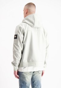 Lichtgrijze hoodie gemaakt van glad materiaal, voorzien van een patch op de linker mouw, met ribgebreide boorden en zoom, gecombineerd met blauwe jeans.