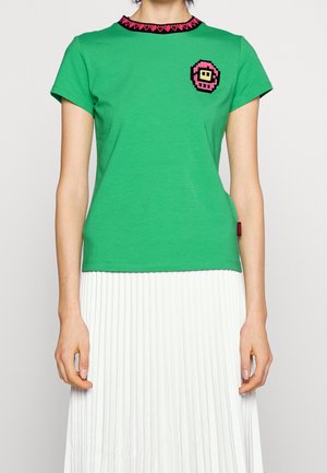 Vrouw draagt een groen t-shirt met roze hartbies op de kraag en een pixel art patch, gecombineerd met een witte plissérok.