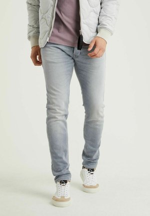 Man wearing grey quilted jacket, mauve shirt, light blue jeans, and white sneakers, standing against a plain background.