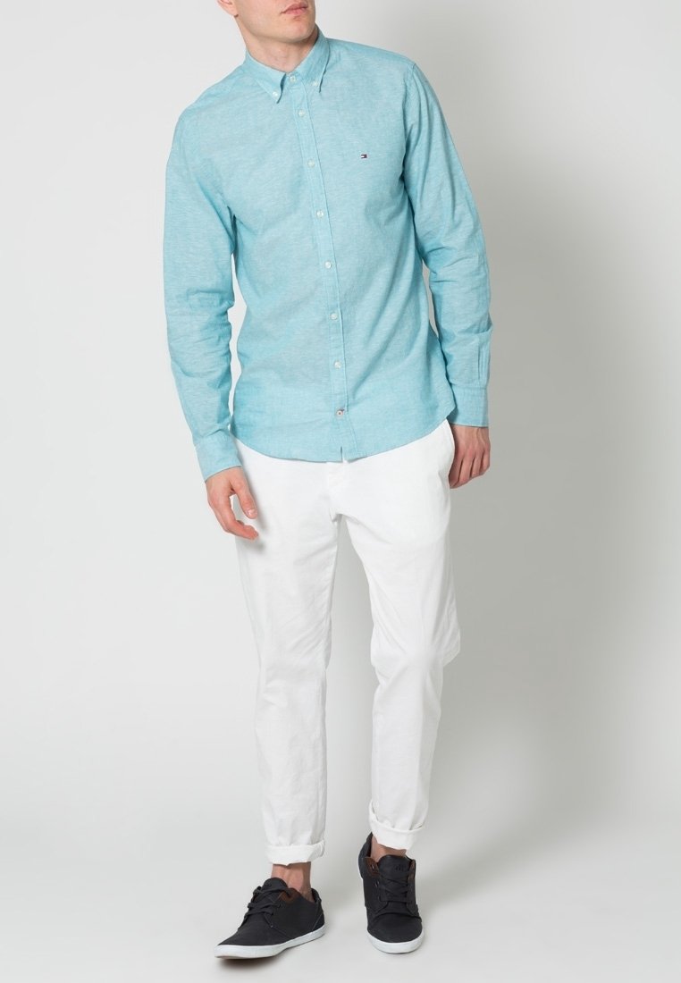 Homme portant une chemise bleu clair à boutons, un pantalon blanc retroussé et des baskets noires, debout sur un fond blanc uni.