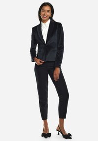 Blazer de terciopelo negro con solapas de satén, combinado con pantalones negros a medida y zapatos de tacón tipo slingback con detalles de lazo; camisa blanca fresca debajo.
