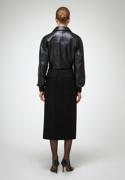 Giubbotto di pelle nera con design accorciato, maniche oversize, polsini a costine; abbinato a una gonna a tubino nera a vita alta e tacchi neri.