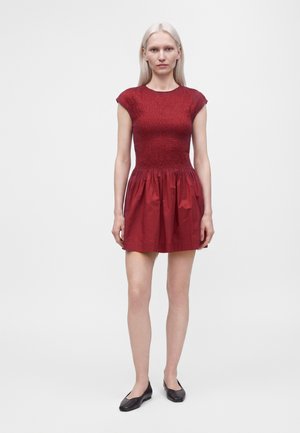Femme aux longs cheveux blonds portant une robe rouge à manches courtes avec un haut ajusté et une jupe froncée, debout devant un fond blanc uni.