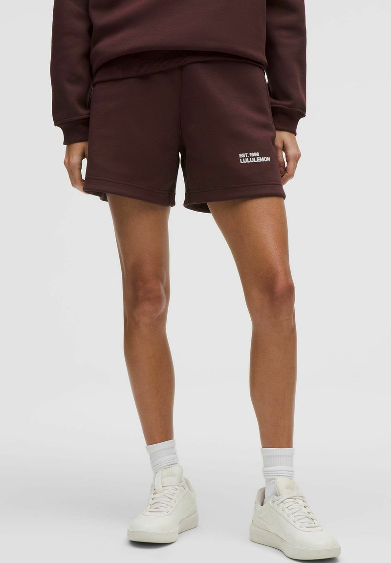 Persoon die donkerbruine korte broek draagt met het "Lululemon"-logo, witte sokken en witte sneakers, staande tegen een effen lichte achtergrond.