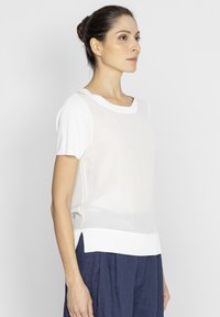 Biała bluzka z krótkim rękawem, wykonana z lekkiego materiału, z zaokrąglonym dekoltem i nieco dłuższym tyłem z bocznymi rozcięciami.