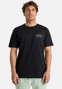 Sort bomulds T-shirt med et lille broderet logo på brystet, rund hals og kortærmet. Modellen har lysegrønne bukser på.