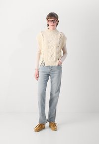 Giovane con capelli corti che indossa occhiali, un maglione senza maniche in maglia color crema sopra una maglietta a maniche lunghe chiara, jeans azzurri e scarpe marroni.