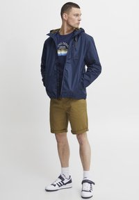 Marine lett jakke med hette, glidelås foran og snor. Kombinert med rullet khaki-shorts og hvite sneakers med marineblå detaljer.