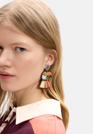 Blondynka nosząca kolorowe kolczyki w kształcie tukana z koralików z frędzlami, jasnoróżowa koszula z kołnierzykiem, patrząca na bok.