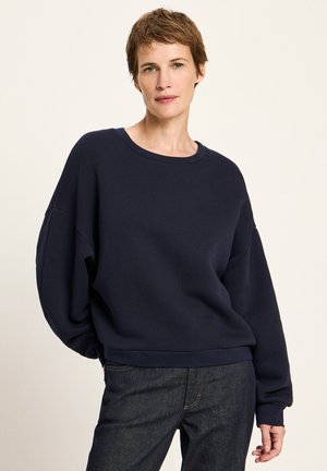 Woman with short brown hair wearing a loose dark navy sweatshirt and dark denim jeans, standing against a plain light background.