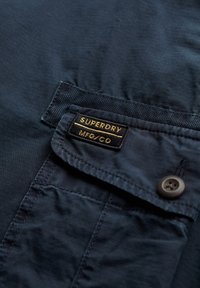 Mørkeblå stof med en subtil ribbet tekstur, der har en sort lap med guld skrift "SUPERDRY MFG/CO" over en knappet lomme.