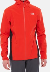 Röd mjukskaljacka med svarta sidopaneler. Har en hel frontdragkedja och justerbar huva. Materialet verkar hållbart och lämpligt för utomhusbruk.