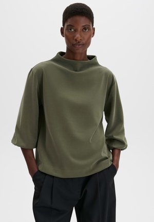 Persona con i capelli corti che indossa una maglia larga verde oliva con maniche al gomito e pantaloni neri, in piedi con le mani nelle tasche.