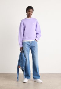 Felpa viola con un piccolo logo, jeans a gamba larga in denim chiaro, sneakers bianche e una giacca di denim sotto il braccio su uno sfondo neutro.