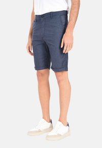 Pantaloni corti blu navy da uomo realizzati in tessuto leggero, con orli arrotolati e chiusura con bottone. Abbinati a sneakers bianche.
