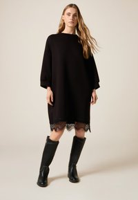 Robe noire surdimensionnée à manches trois-quarts, avec un ourlet en dentelle. Associée à des bottes en cuir noir jusqu'au genou. Texture lisse.