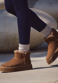 Barna velúr bokacsizmák műszőrme díszítéssel, lapos talppal és dekoratív varrással, szürke bordázott zoknikkal és sötétkék leggingsszel kombinálva.