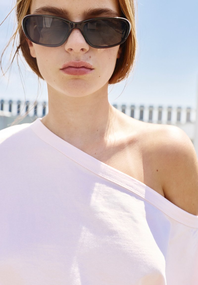 Femme aux cheveux courts portant des lunettes de soleil ovales noires et un haut blanc à épaules dénudées sous un fort soleil en extérieur.