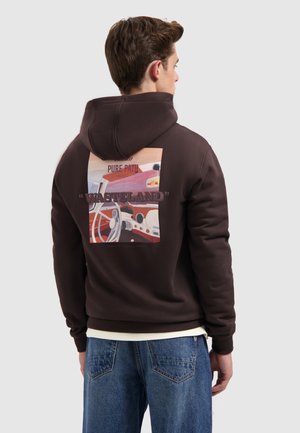 Jongeman in een donkere hoodie met een kleurrijke retro autocomputerprint en de tekst "WASTELAND" op de achterkant, gecombineerd met blauwe jeans.