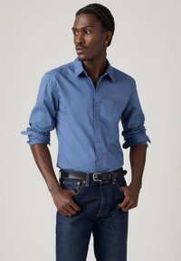 Lichtblauwe lange mouwen overhemd van een gladde katoenen stof, met een button-down kraag en opgerolde mouwen. Gecombineerd met donkere denimbroek en een zwarte riem.