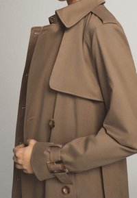 Personne portant un trench-coat marron avec des détails de boutons et de boucles, ajustant l'ouverture frontale du manteau.