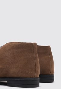 Bottines en suedine marron avec une texture lisse, une forme arrondie et une semelle en caoutchouc noir. Présente un design minimaliste sans lacets visibles.