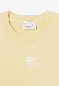 Sudadera unisex de color amarillo claro con cuello redondo. Presenta un logo blanco de Lacoste y el texto "PARÍS" en la parte frontal. Tejido de textura suave.