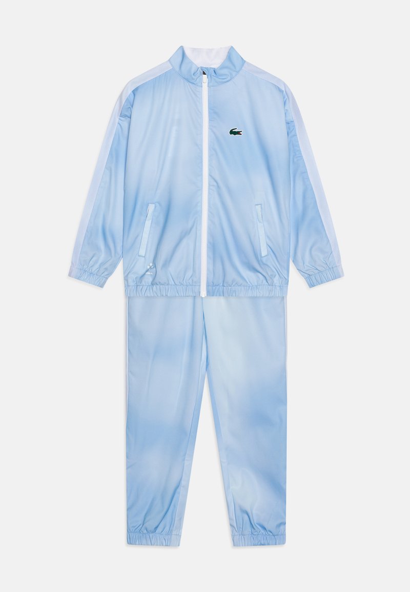 Survêtement bleu clair en polyester brillant. La veste présente un col haut et une fermeture éclair. Le pantalon a des poignets élastiques et des poches.