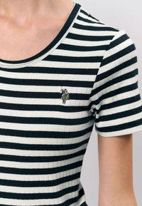 Musta-valkoraidallinen lyhythihainen t-paita, jossa on pyöreä kaula-aukko ja resorikuvio, sekä pieni kirjailtu yksityiskohta rinnassa.