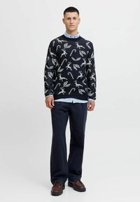 Pull navy avec un motif de renne festif et de canne à sucre, en tissu tricoté texturé, associé à une chemise à col bleu clair et un pantalon foncé.