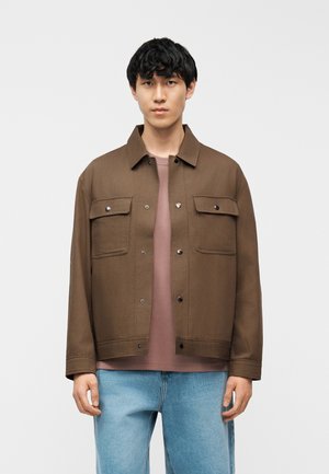 Fiatal férfi előre nézve, barna gombos dzsekit visel mauve színű ing és világoskék farmer felett, egyszínű fehér háttér előtt.