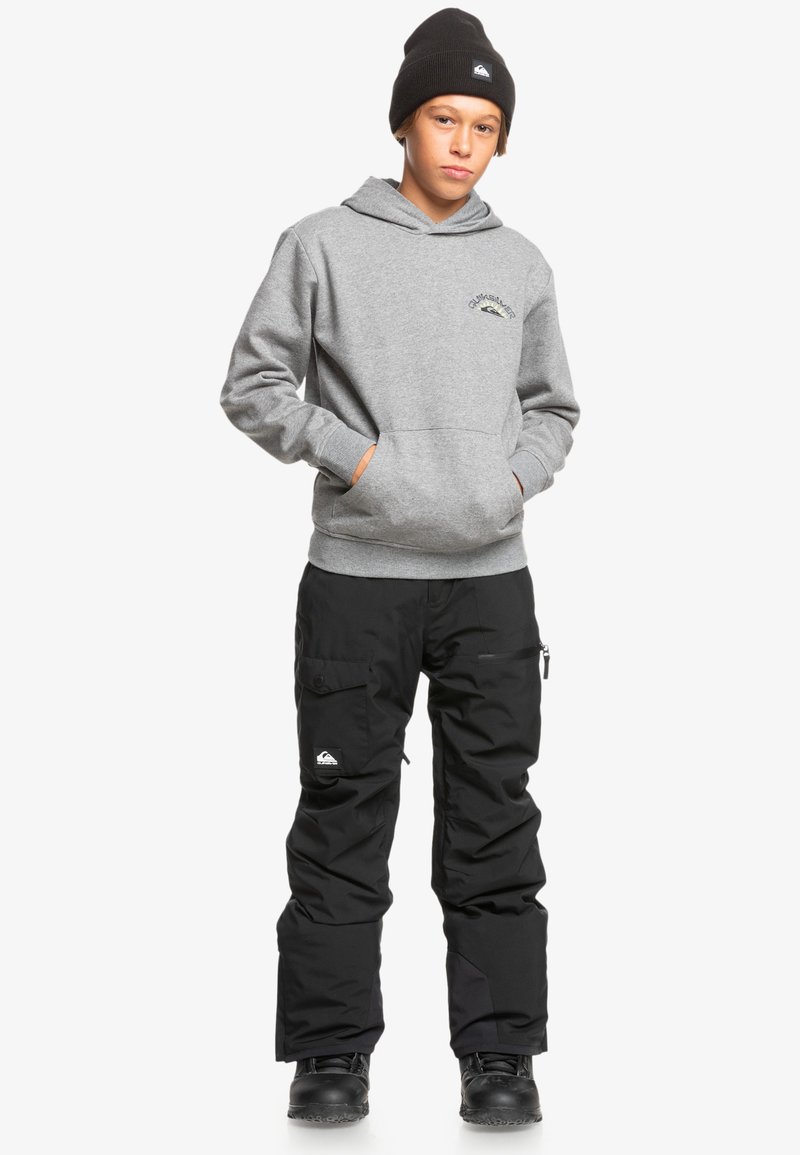 Sudadera gris con un bolsillo frontal y un logo, pantalones de nieve cargo negros con bolsillos laterales, combinados con botas negras y una gorra de lana.