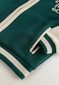 Sudadera verde oscura con puños y rayas color crema, que presenta una textura suave y un detalle de costura visible en los puños.